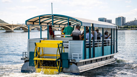 Colorful paddlewheel tour boat with a yellow wheel and covered passenger deck cruising on an urban river, passing an arched bridge and modern city skyline.
