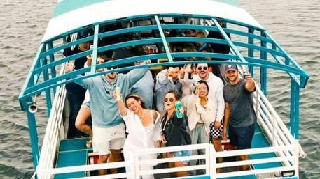 Group of friends on a teal-and-white canopy boat holding drinks and waving on a daytime coastal boat party over calm water