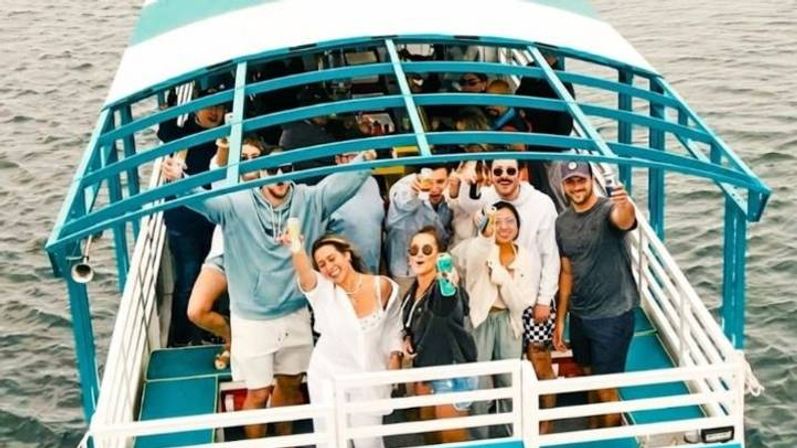 Group of friends on a teal-and-white canopy boat holding drinks and waving on a daytime coastal boat party over calm water