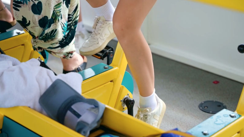 Close-up of two people pedaling a bright yellow stationary exercise bike indoors — white sneakers and socks, tropical-print pants, towel and ankle brace visible.