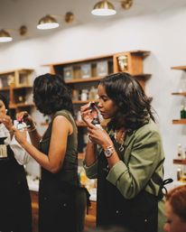 Women in a boutique fragrance workshop sampling perfume from dropper bottles by wooden display shelves