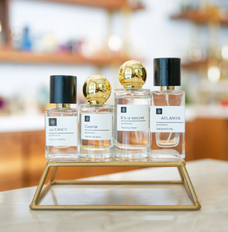 Chic row of four glass perfume bottles with black and gold caps on a brass display stand at a boutique counter, soft blurred shop background