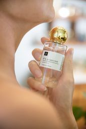 Close-up of a person holding a clear glass perfume bottle with a shiny gold spherical cap and label reading "it's a secret," held close to the neck — elegant fragrance bottle.