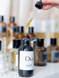 Golden droplet from a glass dropper falling into a black bottle labeled 'oud' on a white vanity, with blurred amber fragrance bottles in the background