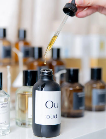 Golden droplet from a glass dropper falling into a black bottle labeled 'oud' on a white vanity, with blurred amber fragrance bottles in the background