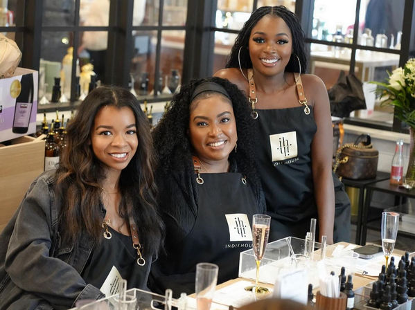 Three smiling women in black aprons at a perfume-making workshop, seated at a table with dropper bottles, pipettes and champagne flutes in a bright beauty studio.