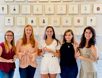 Five smiling women in a fragrance boutique holding perfume bottles, bride-to-be in white with veil in center, framed perfume prints on the wall behind