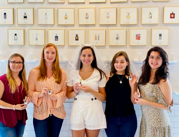 Five smiling women in a fragrance boutique holding perfume bottles, bride-to-be in white with veil in center, framed perfume prints on the wall behind