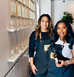 Two smiling women in aprons pose inside a bright boutique scent studio, one holding an amber pour bottle beside shelves of glass sample jars and framed art.