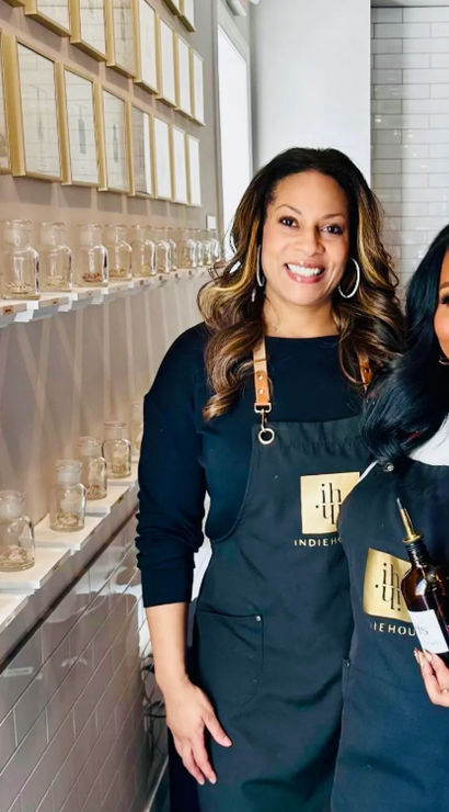 Two smiling women in aprons pose inside a bright boutique scent studio, one holding an amber pour bottle beside shelves of glass sample jars and framed art.