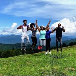 Five people jumping joyfully on a grassy hilltop overlook with rolling blue mountains and a bright cloudy sky