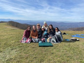 Seven friends wrapped in woven blankets sitting on a grassy mountaintop meadow, smiling with layered mountain ridges and a blue sky backdrop.