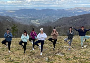 Seven people in winter hats and sunglasses striking yoga tree pose on a grassy mountaintop overlook with a scenic valley and distant blue mountains under a cloudy sky