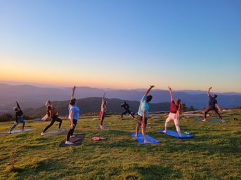 Outdoor sunset yoga class on a grassy mountain ridge — group stretching on mats against a colorful sky and layered blue mountain panorama.