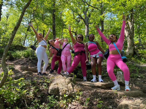 Group of six friends in bright pink activewear cheering with arms raised on a sunlit forest hiking trail among green trees and rocky steps
