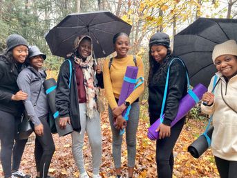 Smiling women holding rolled yoga mats and umbrellas on a rainy autumn woodland trail covered in colorful fall leaves