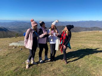 Four friends in winter beanies and jackets toasting with glasses on a grassy mountain overlook, scenic blue ridges and clear sky stretching behind them.