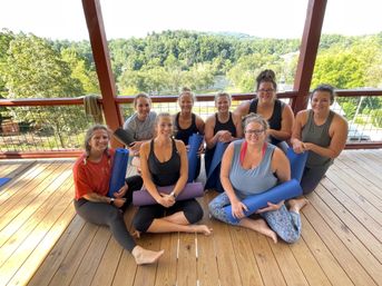 Group of smiling women holding yoga mats on a sunny covered wooden deck with a river and green forested hills in the background — outdoor yoga retreat on a riverfront overlook.