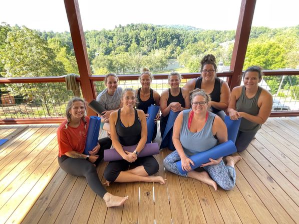 Group of smiling women holding yoga mats on a sunny covered wooden deck with a river and green forested hills in the background — outdoor yoga retreat on a riverfront overlook.