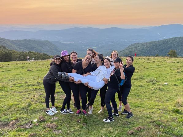 Friends on a grassy mountaintop at sunset, laughing as they lift one friend horizontally, with rolling green hills and layered blue mountains fading into the distance.