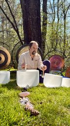 Person seated in a sunlit woodland clearing playing crystal singing bowls with mallets, surrounded by large gongs and a line of rose quartz and crystals on the green grass — outdoor sound bath scene.