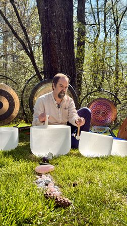 Person seated in a sunlit woodland clearing playing crystal singing bowls with mallets, surrounded by large gongs and a line of rose quartz and crystals on the green grass — outdoor sound bath scene.