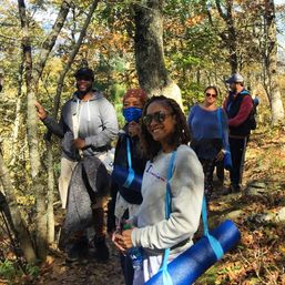 Smiling group on a sunlit autumn forest hike, carrying rolled blue yoga mats and water bottles along a leaf-covered wooded trail