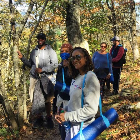 Smiling group on a sunlit autumn forest hike, carrying rolled blue yoga mats and water bottles along a leaf-covered wooded trail