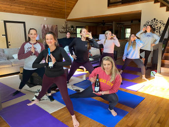 Group of women practicing yoga on mats while sipping wine in a sunlit rustic cabin living room with wood-paneled ceiling and cozy seating
