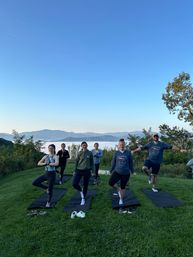 Six people practicing tree pose on yoga mats during a morning outdoor yoga session on a grassy hilltop overlooking a fog-filled valley and blue mountain ridges under a clear sky.