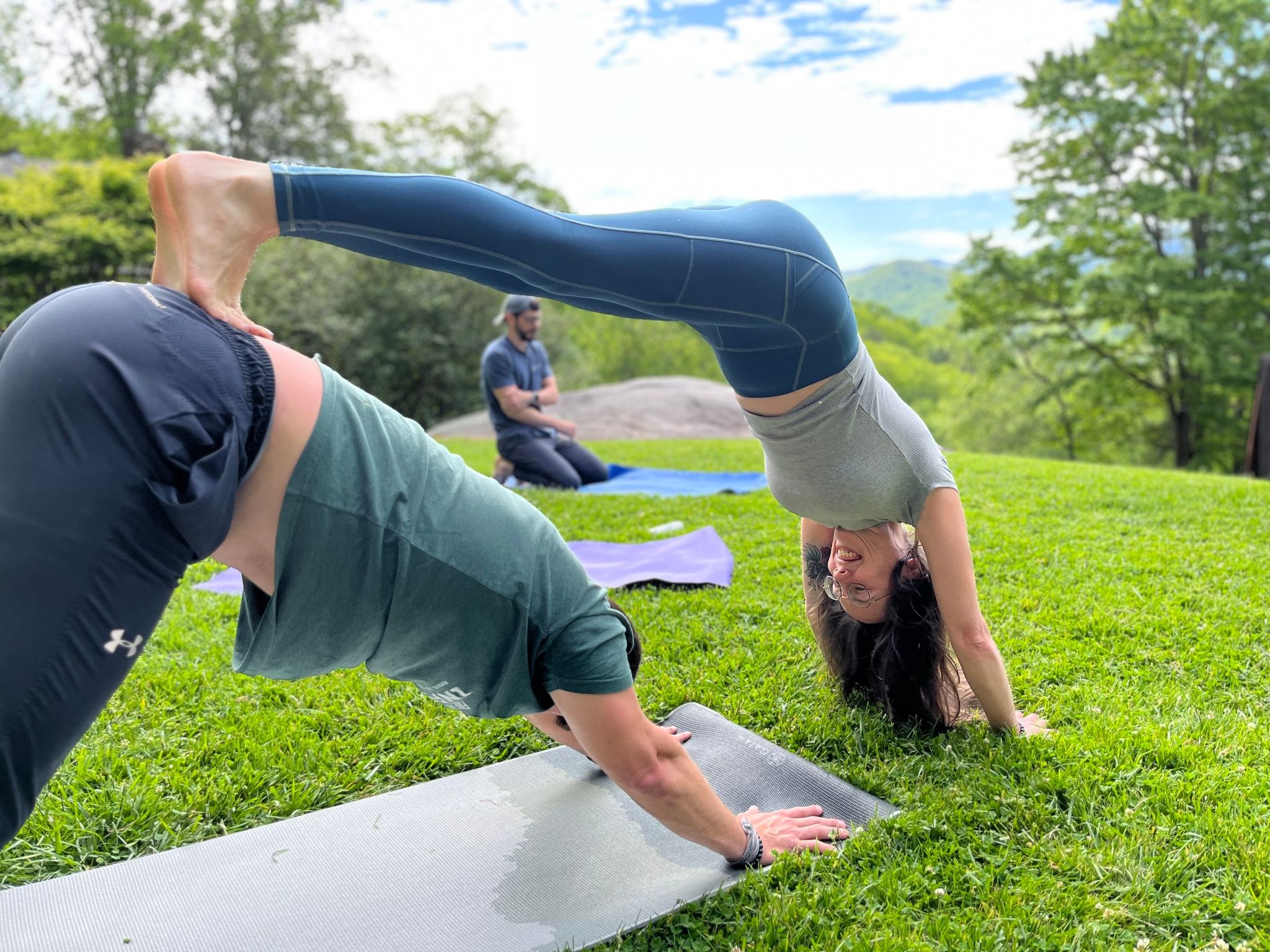 Outdoor partner yoga on a green hillside with mountain views: one person in downward dog supports a partner balancing in a playful inverted backbend, yoga mats and trees in the background.
