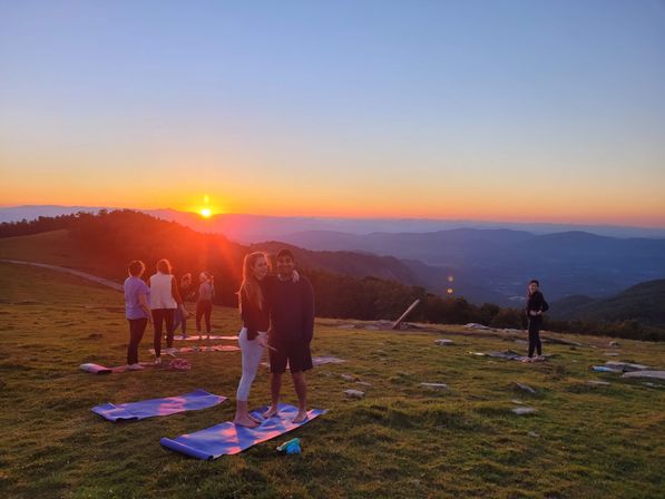 Scenic sunset yoga on a grassy mountaintop meadow — a small group on colorful mats silhouetted against a warm golden-orange sky and layered blue hills.