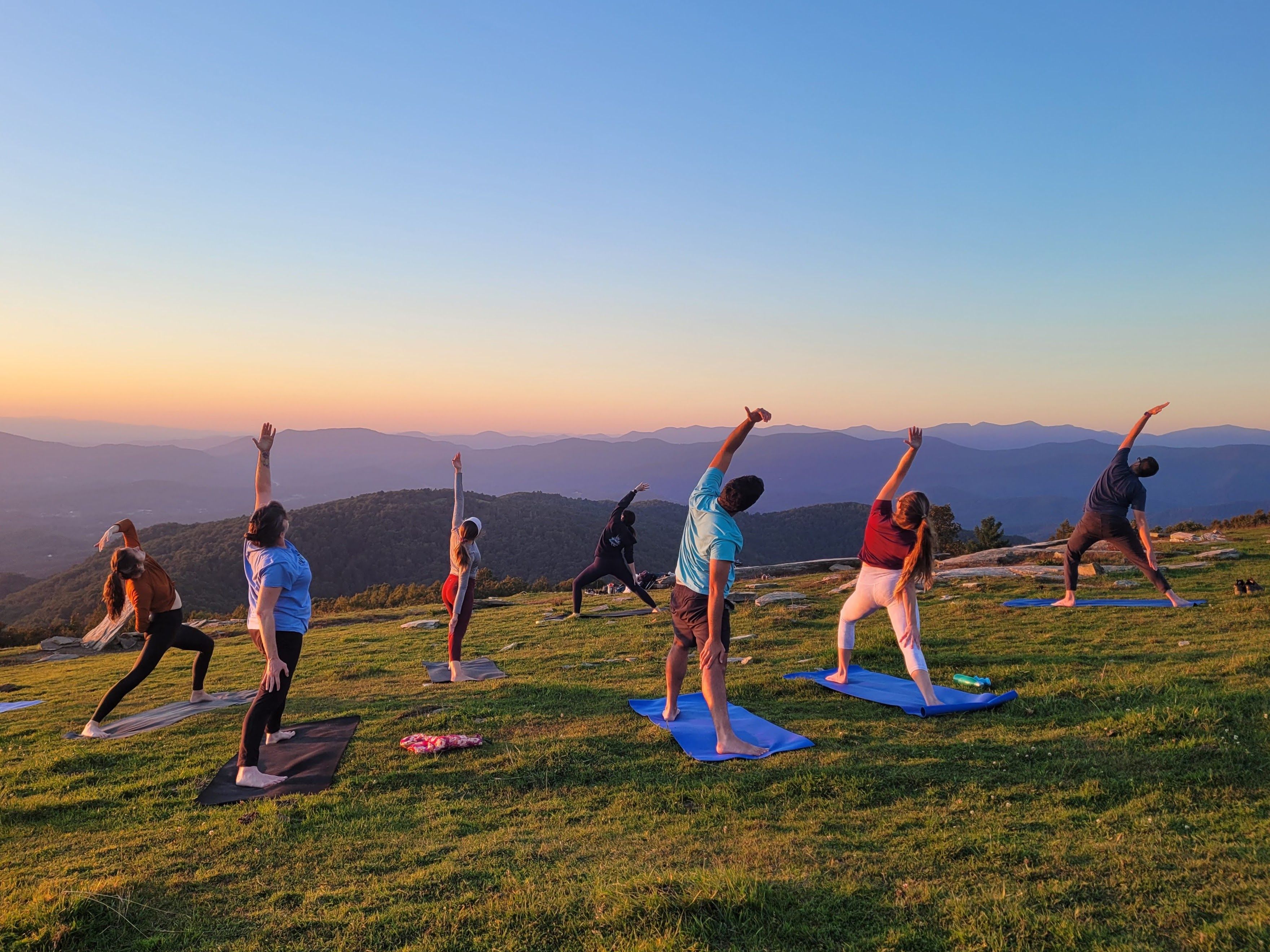 Group sunset yoga on a grassy mountaintop — people on mats stretching toward the sky with layered blue mountain ridges and a colorful sunset sky in the background.