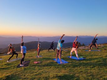 Group sunset yoga on a grassy mountaintop — people on mats stretching toward the sky with layered blue mountain ridges and a colorful sunset sky in the background.