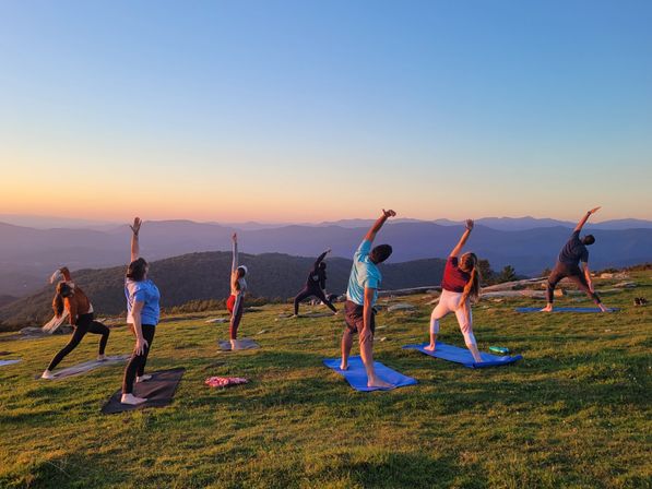 Group sunset yoga on a grassy mountaintop — people on mats stretching toward the sky with layered blue mountain ridges and a colorful sunset sky in the background.