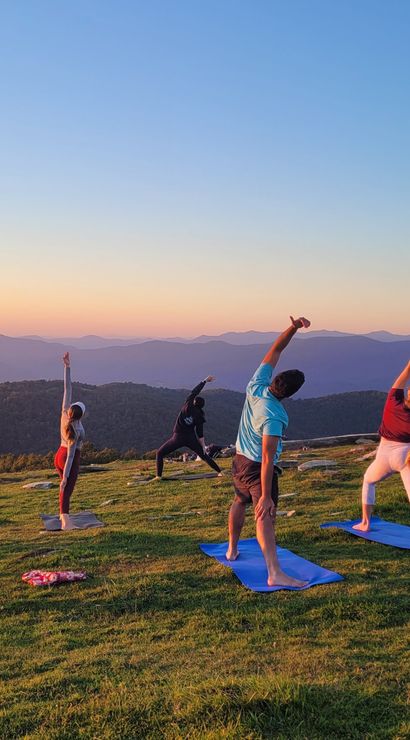 Group sunset yoga on a grassy mountaintop — people on mats stretching toward the sky with layered blue mountain ridges and a colorful sunset sky in the background.