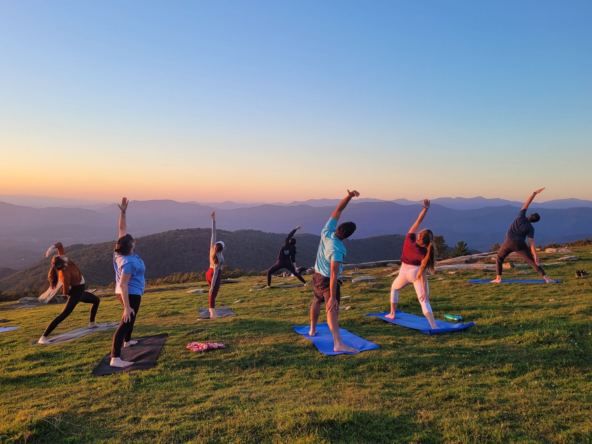 Group sunset yoga on a grassy mountaintop — people on mats stretching toward the sky with layered blue mountain ridges and a colorful sunset sky in the background.