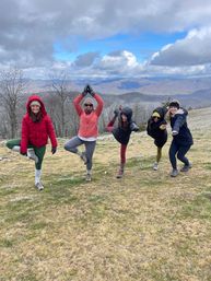 Five women in winter jackets and hats striking playful yoga poses on a grassy mountain overlook during a chilly hike, scenic rolling ridges and dramatic cloudy blue-gray sky in the background