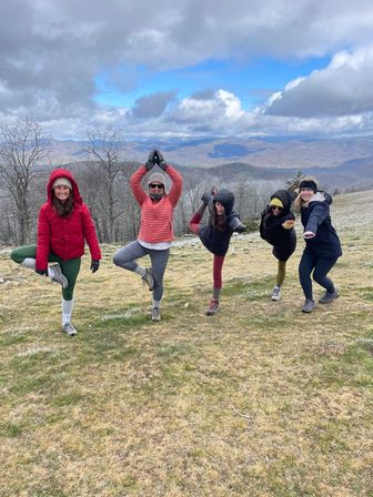 Five women in winter jackets and hats striking playful yoga poses on a grassy mountain overlook during a chilly hike, scenic rolling ridges and dramatic cloudy blue-gray sky in the background