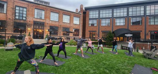 Group practicing outdoor yoga on mats in warrior II pose on a grassy urban courtyard, with a large red-brick industrial-style hotel building and glowing windows in the background.
