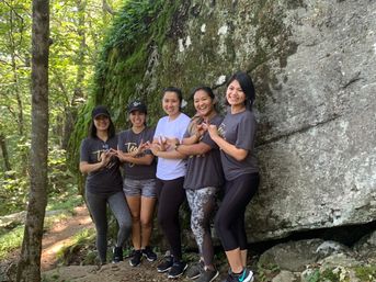 Five women hikers in casual activewear smiling and making hand signs while posing on a sunlit forest trail beside a large moss-covered boulder.