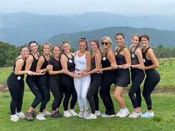 Group of women in black activewear and one in a white sash posing arm-in-arm on a grassy mountain overlook with layered blue ridges — outdoor bachelorette hike and fitness photo.