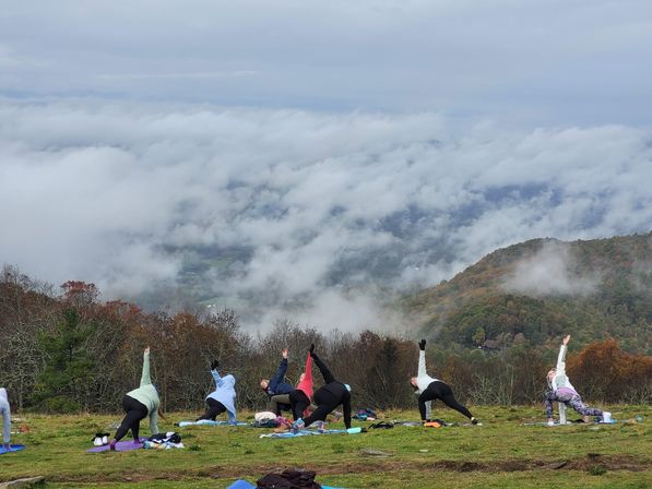 Group outdoor yoga on a grassy mountaintop, people stretching on mats with a foggy valley and colorful autumn hills drifting through low clouds in the background.