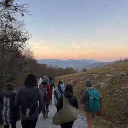 Group of hikers with backpacks walking a gravel mountain trail at sunset, pastel sky and layered blue ridges in the distance.