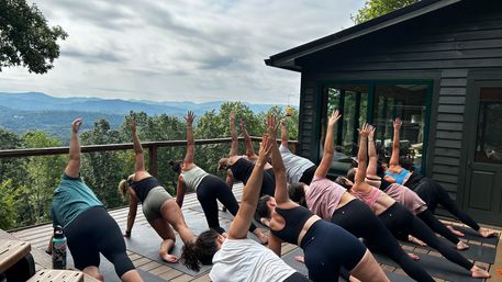 Group outdoor yoga class on a wooden mountain deck, participants in side-plank with raised arms overlooking forested ridges and a cloudy sky