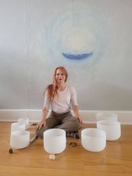 Red-haired woman seated cross-legged on a wooden floor in a bright meditation room, surrounded by white crystal singing bowls in a semicircle with a soft blue crescent mural on the wall — sound bath session.