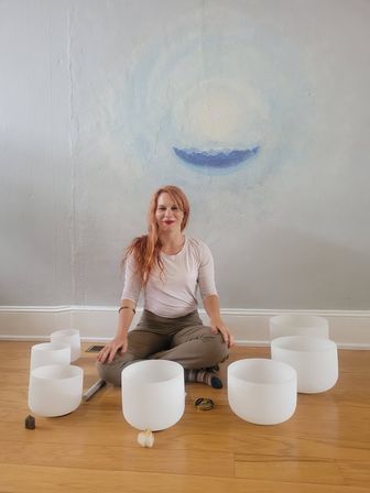 Red-haired woman seated cross-legged on a wooden floor in a bright meditation room, surrounded by white crystal singing bowls in a semicircle with a soft blue crescent mural on the wall — sound bath session.