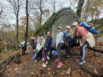 Smiling group of hikers posing on a leaf-strewn forest trail beside a large moss-covered boulder in autumn, carrying rolled mats and backpacks