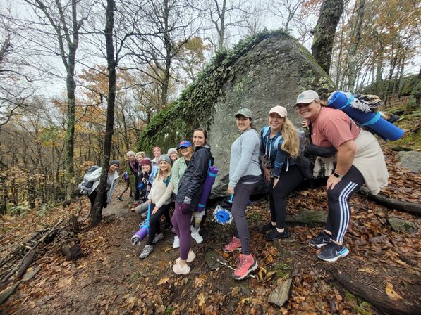 Smiling group of hikers posing on a leaf-strewn forest trail beside a large moss-covered boulder in autumn, carrying rolled mats and backpacks