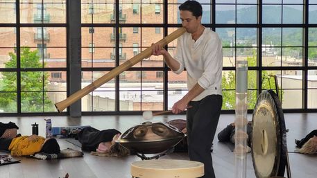 Musician plays a long wooden didgeridoo and taps a handpan and gong during a sound-healing session in a bright urban loft studio with floor-to-ceiling windows and people resting on mats.